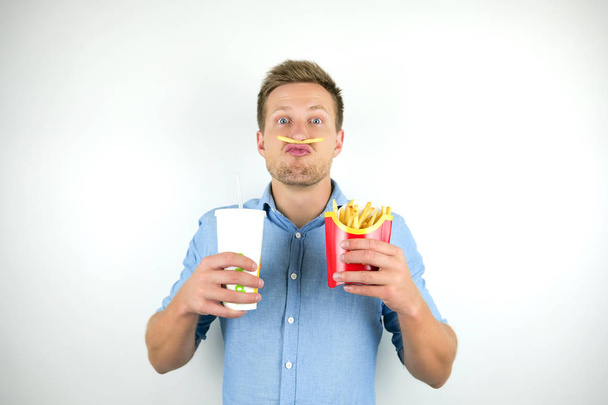 young man standing with one french fries under his nose and holds soda in papper cup and fries from fast food restaurant looks funny on isolated white background - Photo, Image