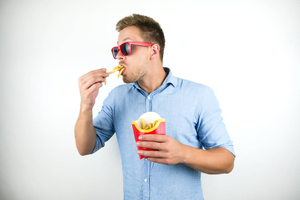 young man in red eyeglasses eats french fries from fast food restaurant looks funny on isolated white background - Foto, imagen