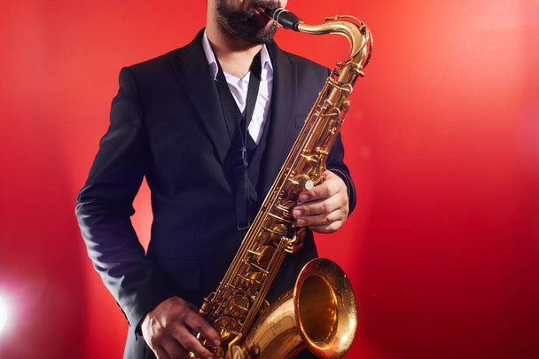 Portrait of professional musician saxophonist man in  suit plays jazz music on saxophone, red background in a photo studio - Photo, Image