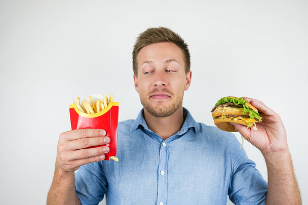 young handsome man holding fries and burger from fast food restaurant looks suspicious on isolated white background - Фото, изображение