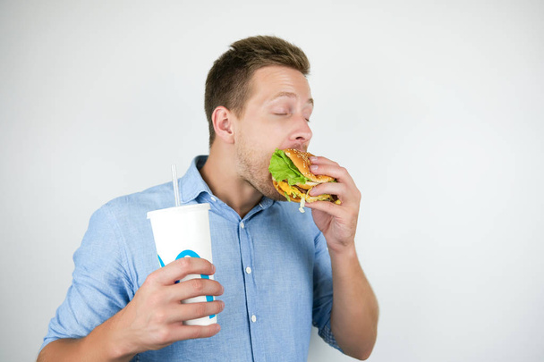 young handsome man holding soda drink in paper cup and biting burger from fast food restaurant looks hungry on isolated white background - Fotografie, Obrázek