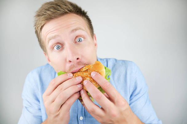 Молодой красивый мужчина, кусающий бургер из ресторана быстрого питания выглядит голодным на изолированном белом фоне
 - Фото, изображение