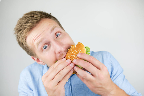 young handsome man eats fresh tasty burger from fast food restaurant looks hungry on isolated white background - Foto, Imagen