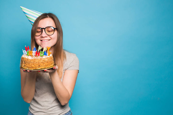 Charming merry crazy young girl student in congratulatory paper hat holding a happy birthday cake in her hands standing against a blue background. Advertising space. - Фото, изображение