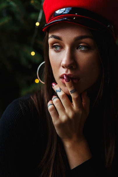 Portrait of beautiful woman touching lips in elegant red cap on background of Christmas tree and looking aside. Christmas glamour photo. Happy new year concept. Copy space on left side - Φωτογραφία, εικόνα