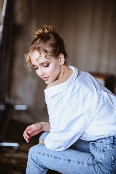 Mujer rubia joven con una camisa blanca casual y jeans en el interior de un loft, retrato de moda de cerca
 - Foto, Imagen