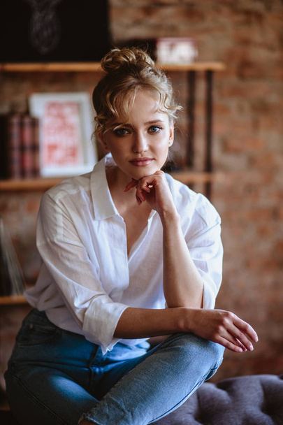 Young blonde woman in a casual white shirt and jeans in a loft interior, close up fashion portrait - Fotó, kép