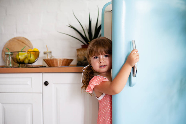 Ένα κοριτσάκι ανοίγει την πόρτα του ψυγείου. Όμορφη κουζίνα. - Φωτογραφία, εικόνα