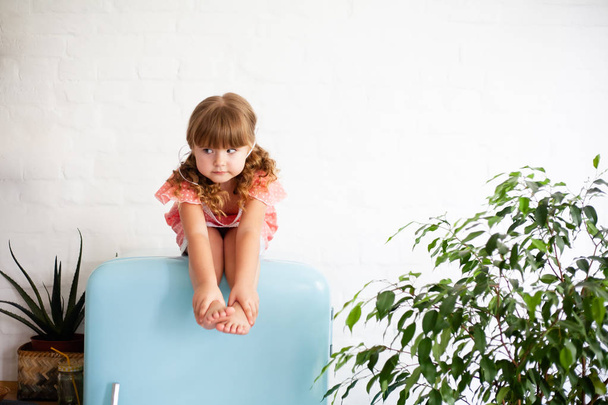 Little girl is sitting on the fridge. Sits on a beautiful retro refrigerator of blue color, stylish interior. - Foto, Imagem