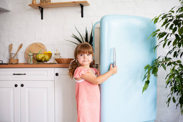A little girl opens the door of the refrigerator. Beautiful kitchen. - Photo, Image