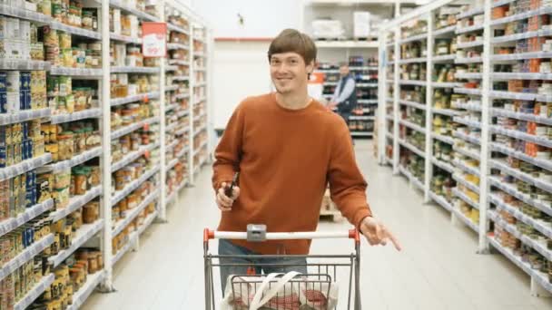 szép fickó egy telefonnal, és egy élelmiszer-kosár táncolnak a szupermarketben - Felvétel, videó