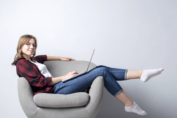 Młoda dziewczyna z laptopem siedzi na miękkim wygodnym fotelu, i sfrezowanie, Kobieta za pomocą komputera przed białą pustą ścianę, ona Freelancing i drukowanie tekstu, Kopiuj przestrzeń - Zdjęcie, obraz