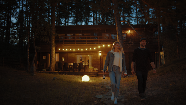 Happy Young Man and Woman Walk From a Cozy Country House Together. House has Warm Lights Hanging Above the Terrace. Romantic Summer Evening Atmosphere. Cottage is Situated in a Pine Forest. - Footage, Video
