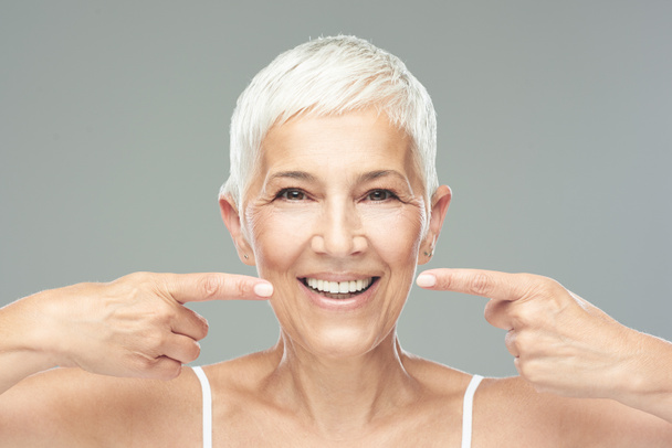 Beautiful Caucasian  smiling senior woman with short grey hair pointing at her teeth and looking at camera. Beauty photography. - Фото, изображение