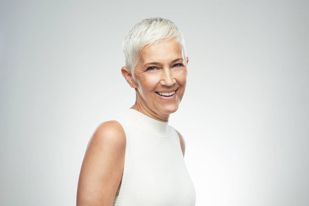 Bella donna anziana sorridente con i capelli corti grigi in posa di fronte a sfondo grigio. Foto di bellezza
. - Foto, immagini