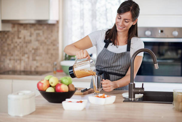 Beautiful smiling Caucasian woman in apron standing in kitchen and pouring fresh smoothie into glass. - 写真・画像