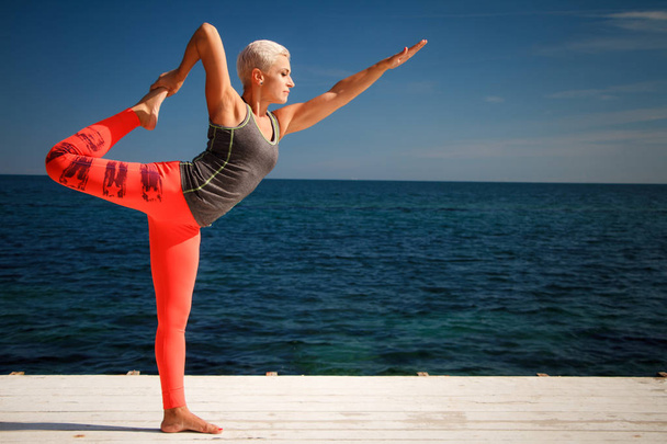 Взрослая блондинка с короткой стрижкой практикует йогу на пирсе на фоне моря и голубого неба
 - Фото, изображение