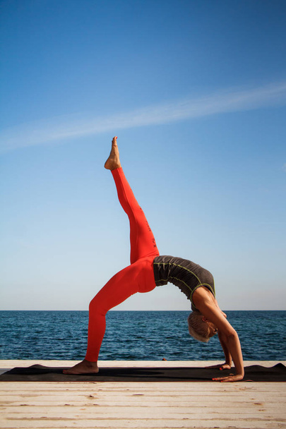 Adult blond woman with short haircut practices yoga on the pier against the background of the sea and blue sky - Photo, Image