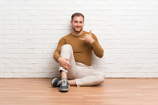 Homem loiro sentado no chão fazendo gesto de telefone
 - Foto, Imagem