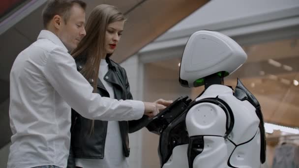 Ein Mann und eine Frau in der Mall interagieren mit einem Roboterberater, indem sie auf den Bildschirm tippen und lächeln. Cyborg-Androide helfen Menschen. - Filmmaterial, Video