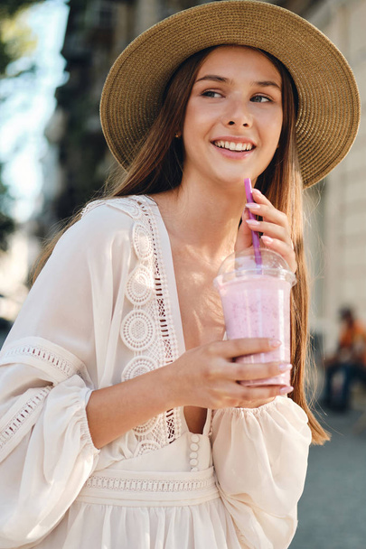 Young attractive smiling woman in white dress and hat holding smoothie to go in hands joyfully looking aside standing on cozy city street alone - Photo, Image