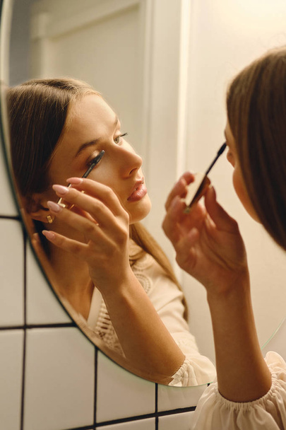 Close up giovane donna attraente in abito bianco in piedi vicino specchio sognante applicazione mascara nero in bagno
 - Foto, immagini