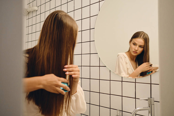 junge hinreißende Frau in weißem Kleid streicht sich nachdenklich die Haare in der Nähe des Spiegels im Badezimmer - Foto, Bild