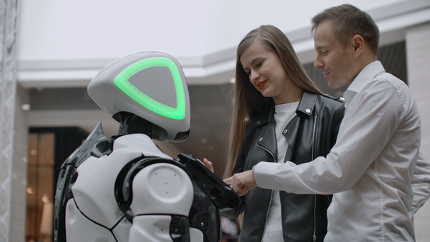 Мужчина и женщина в торговом центре взаимодействуют с консультантом-роботом, нажимая на экран и улыбаясь. Cyborg Android помогает людям
. - Кадры, видео