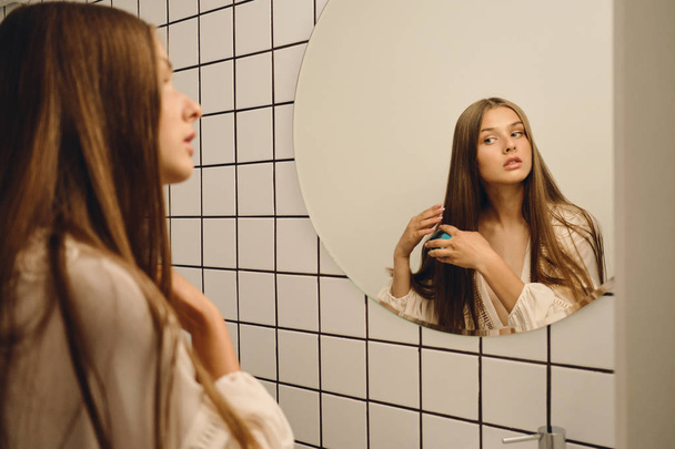 Nuori kaunis nainen valkoisessa mekossa unelmoi peilistä harjaamalla hiuksiaan kylpyhuoneessa
 - Valokuva, kuva