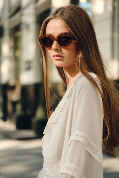 Young beautiful woman in sunglasses and white dress thoughtfully looking in camera standing on cozy city street alone - Foto, afbeelding