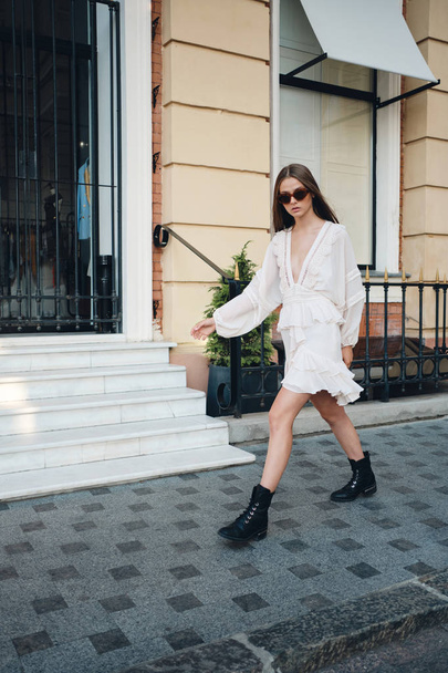 Jonge aantrekkelijke serieuze vrouw in zonnebril, mooie witte jurk en zwarte laarzen doordacht op zoek in camera wandelen op City Street  - Foto, afbeelding