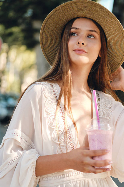Young beautiful woman in white dress and hat holding smoothie to go in hand dreamily looking aside standing on cozy city street  - Photo, Image