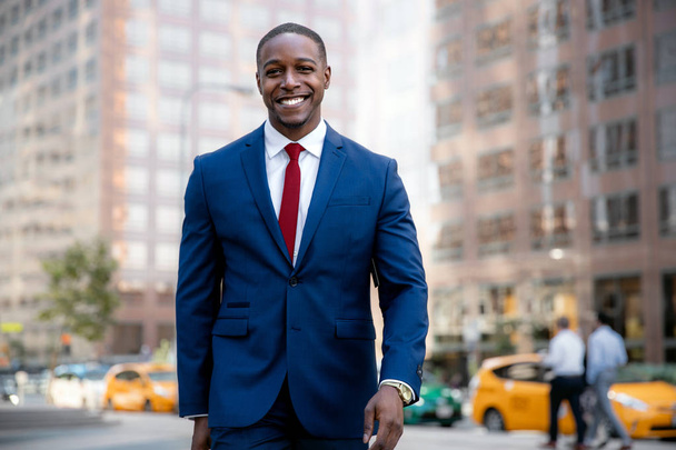 Professionista d'affari afroamericano in giacca e cravatta, sorridente mentre cammina verso l'ufficio sul posto di lavoro nel distretto finanziario, sfondo urbano
 - Foto, immagini