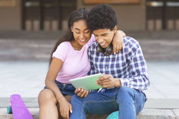 Dark-skinned tieners kijken naar video op digitale Tablet zittend buitenshuis - Foto, afbeelding