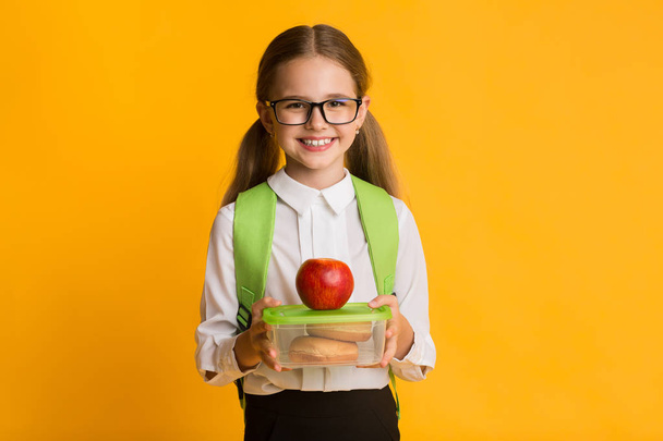 Cute Schoolgirl Holding Lunch Box Over Yellow Studio Background - Foto, Imagen
