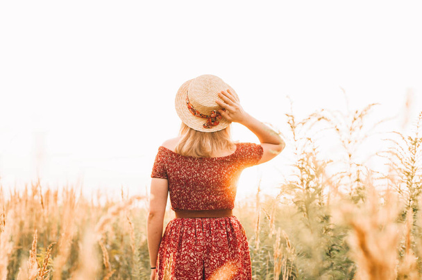 Beautiful girl in red dress in yellow walking through field, sunset time. golden hour. village scenery - Photo, Image