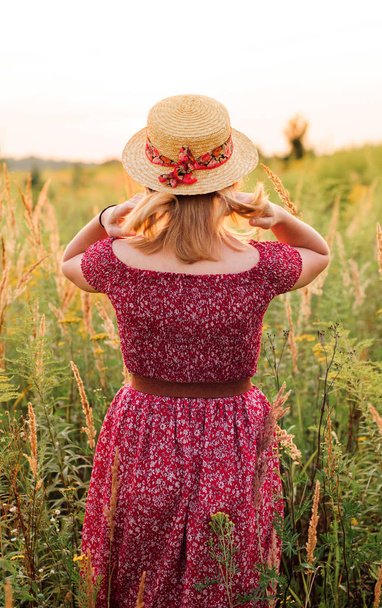 Beautiful girl in red dress in yellow walking through field, sunset time. golden hour. village scenery - Photo, image