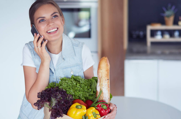 Smiling woman with mobile phone holding shopping bag in kitchen - Foto, imagen