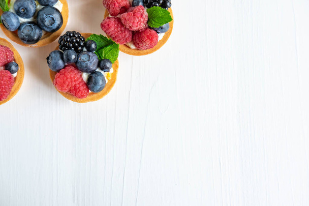 Photo pattern with tartlets on a white wooden background. Cakes with berries of marina, blueberry, blueberry with mint leaves. Photo patern with sweets top view - Photo, image