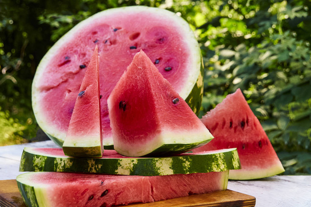 Fresh ripe striped sliced watermelon on a wooden old table, against the background of green leaves, outdoors. - Photo, Image