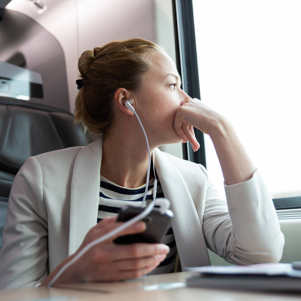 Στοχαστική επιχειρηματίας ακούγοντας podcast στο κινητό τηλέφωνο, ενώ ταξιδεύετε με τρένο. - Φωτογραφία, εικόνα
