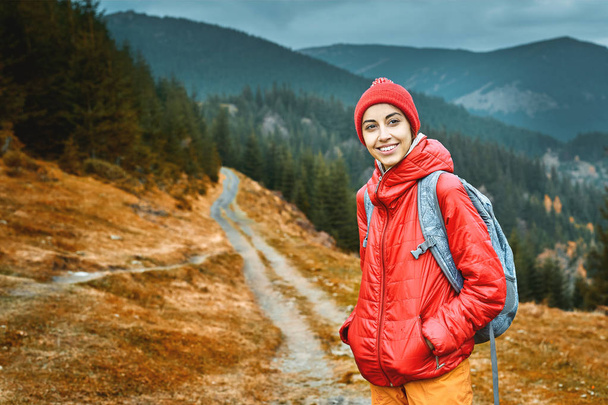 woman hiker with backpack, wearing in red jacket and orange pants, standing on the mountains background - Фото, изображение