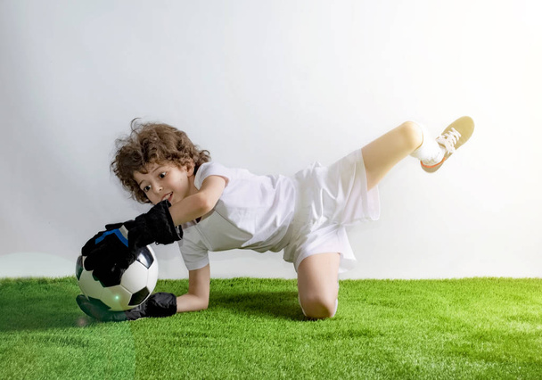 Goalkeeper lying on the grass catches a ball. Excited little toddler boy playing football on soccer field against light background. Active childhood and sports passion concept. Save space - Foto, Imagen