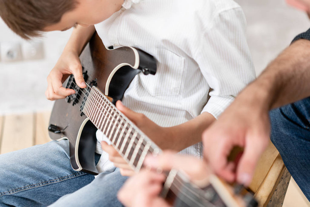 Young boy moves his hand along guitar strings while his father helps boy to take chords - Valokuva, kuva
