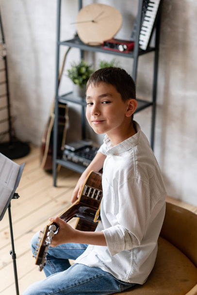 Young boy in casual clothing sits on chair with acoustic guitar in hands and posing - Fotografie, Obrázek