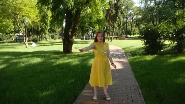 Vöröshajú lány fúj szappanbuborékok a parkban. sárga ruha mosolygott és nevetve. nyár és boldogság  - Felvétel, videó