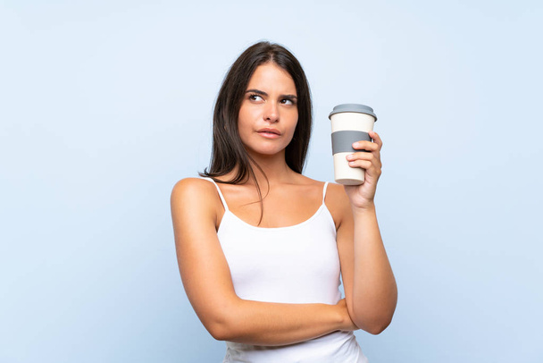 Mujer joven sosteniendo un café para llevar sobre fondo azul aislado mirando hacia arriba mientras sonríe
 - Foto, imagen