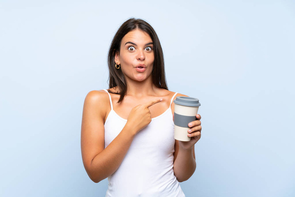 Молодая женщина, держащая забрать кофе на изолированном синем фоне удивлен и указывая стороны
 - Фото, изображение