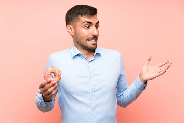 Νέος άνθρωπος κρατώντας ένα ντόνατ πάνω από απομονωμένο ροζ φόντο με έκπληξη έκφραση του προσώπου - Φωτογραφία, εικόνα