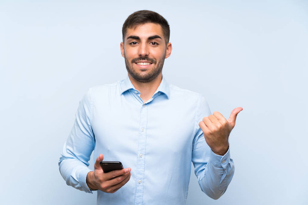 Młody przystojny człowiek z jego telefon komórkowy przez izolowany niebieski ściana wskazując na bok, aby przedstawić produkt - Zdjęcie, obraz